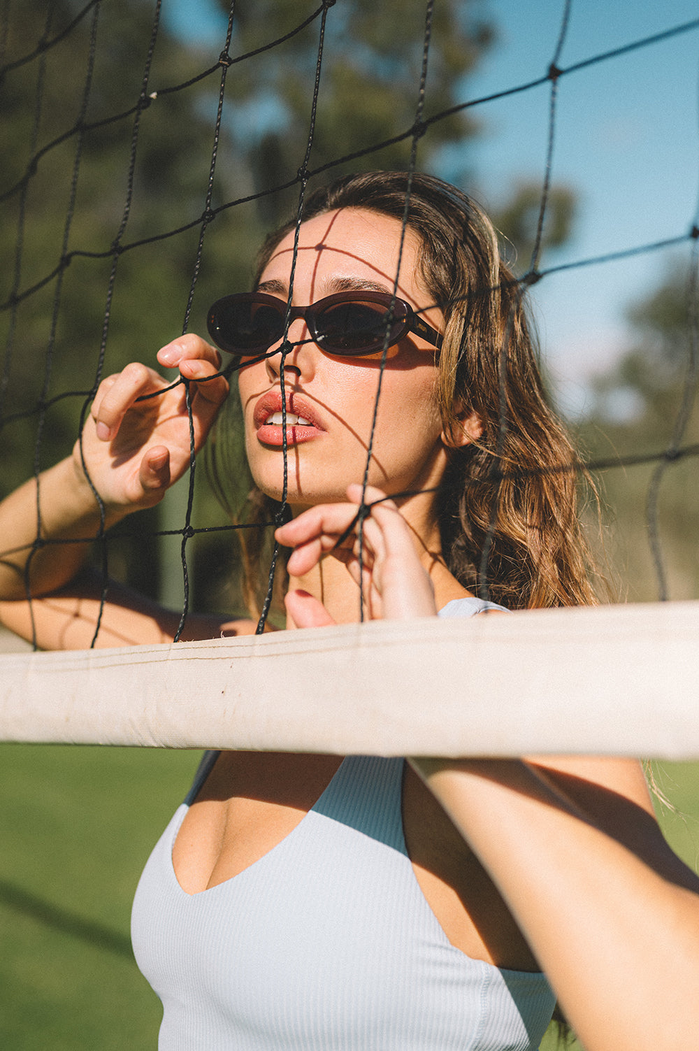 Woman adjusting brown oval sunglasses behind a volleyball net with a blurred natural background