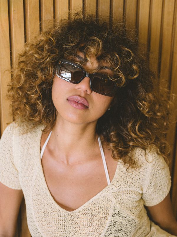 Woman with curly hair wearing brown rectangle sunglasses and a beige top against a wooden background