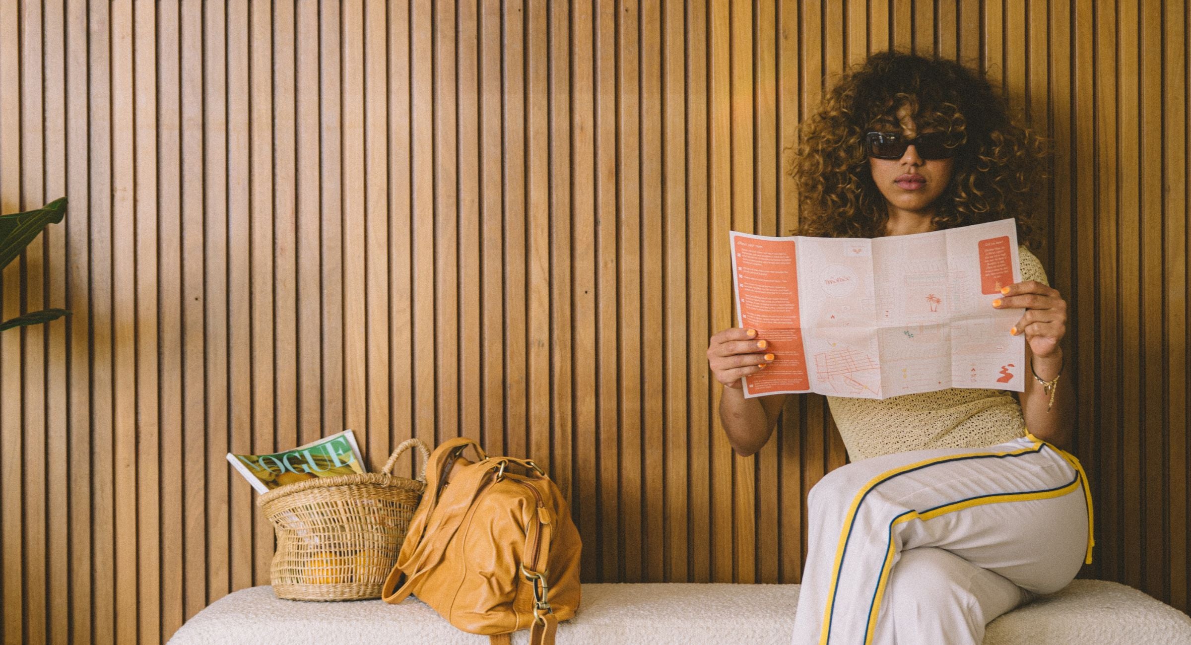 Woman wearing brown sunglasses reading a map in a room with wooden walls and a leather bag and basket on a chair.