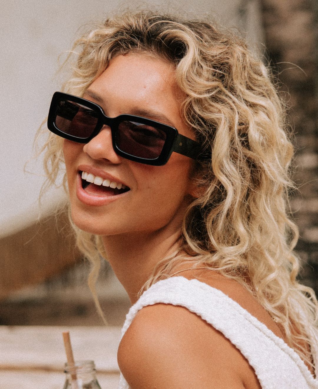 Person with curly blonde hair wearing black rectangle women's sunglasses and a white top.